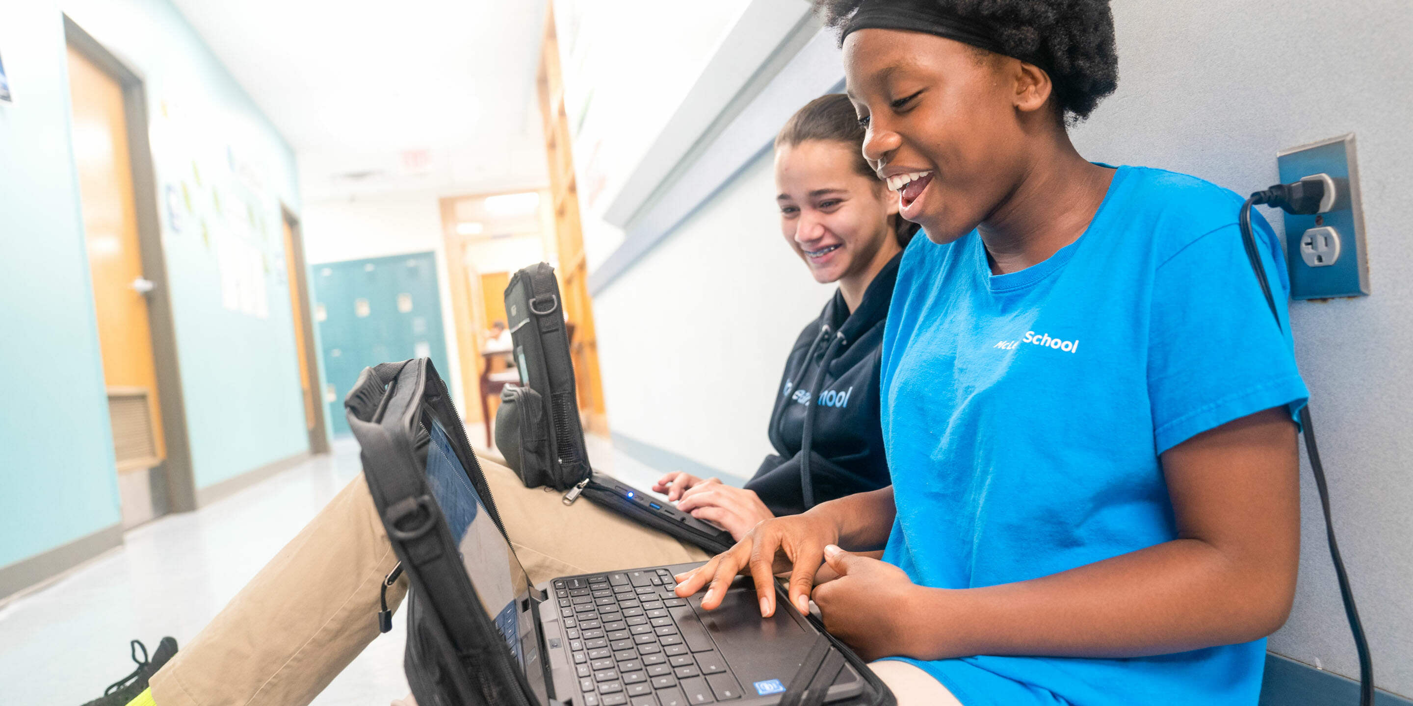 Two girls sit in school hallway with laptops