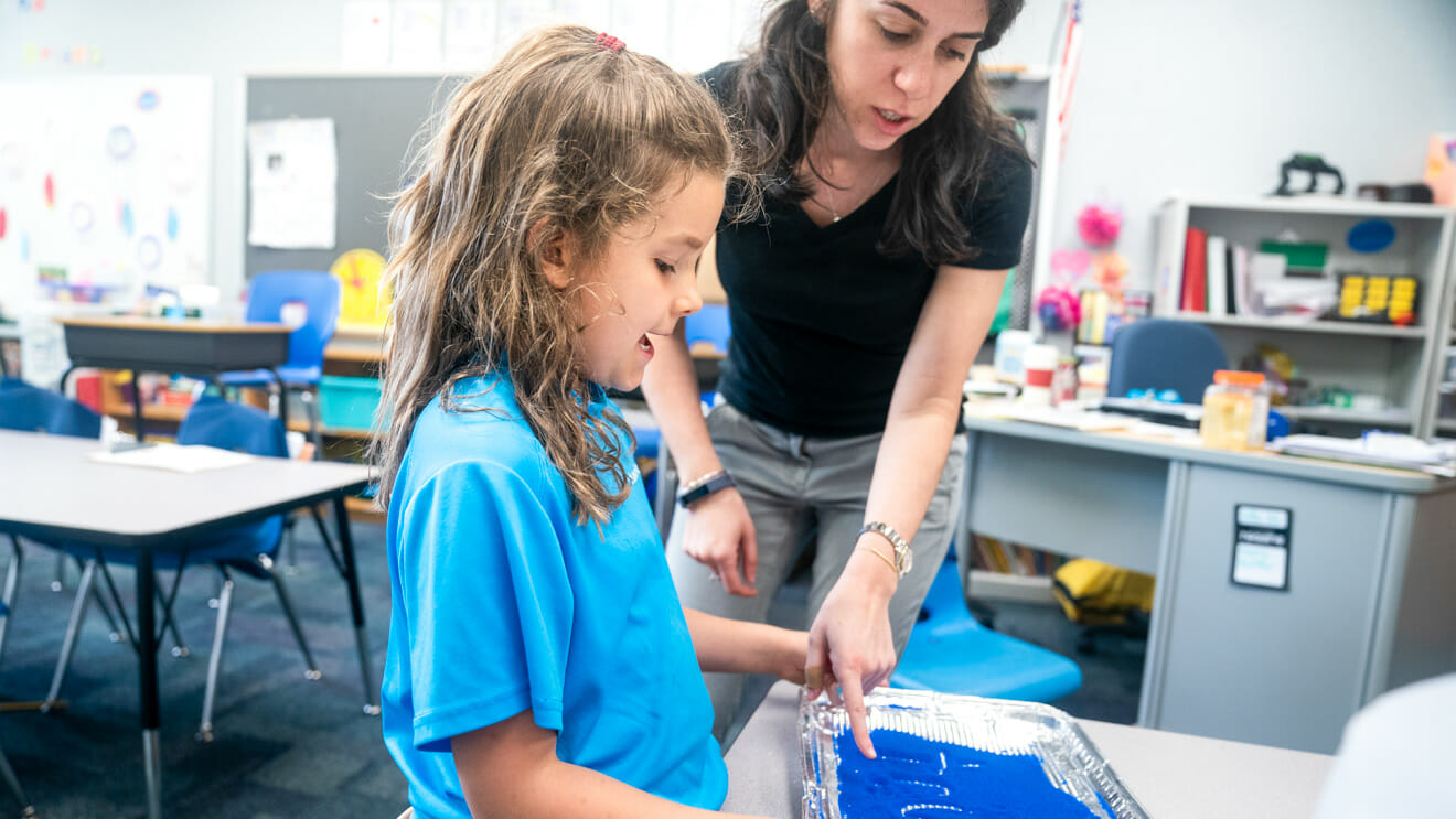 Teacher helps girl write in sand tray