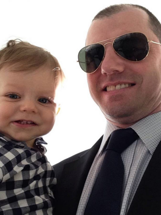 Mark Reyman, Middle School Literature Teacher at McLean School, smiles at camera with a suit and sunglasses with his child