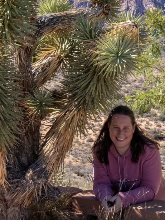 Karen Rappaport, Learning Commons Librarian at McLean School, smiles at camera while kneeling next to a tree