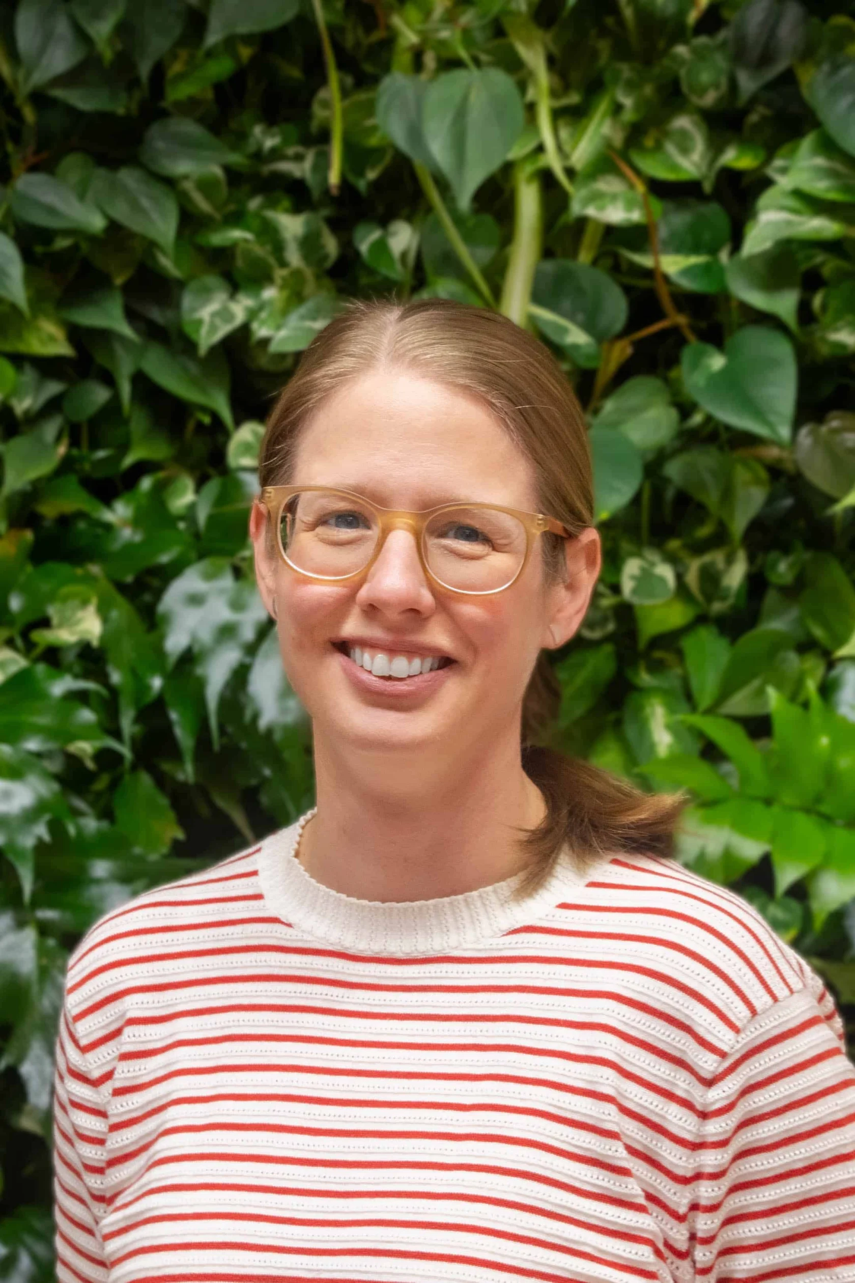 Mclean-Natalie-Stapert A person with glasses and a striped sweater stands in front of a green leafy background, smiling at the camera.