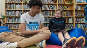 Two students sit on the floor of a library, reading together and smiling. Bookshelves filled with books are visible in the background.