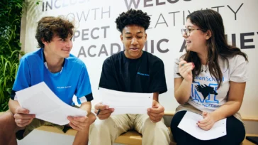 Three students sit on a bench indoors, holding papers and talking, with large text on the wall behind them featuring words like “welcoming” and “academic.”.