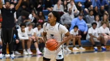 A basketball player in a white uniform holds the ball, preparing to make a move during a game in a gym with spectators watching from the bleachers.