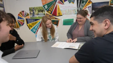 Four students and an adult sit around a table discussing documents, with college pennants displayed on the wall behind them.