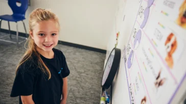 A young girl with long blonde hair stands by a whiteboard with images and words, smiling at the camera in a classroom setting.