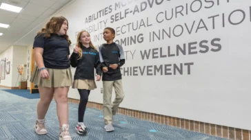 Three students walk together in a school hallway beside a wall with motivational words such as "SELF-ADVOCACY" and "ACHIEVEMENT" displayed in large letters.