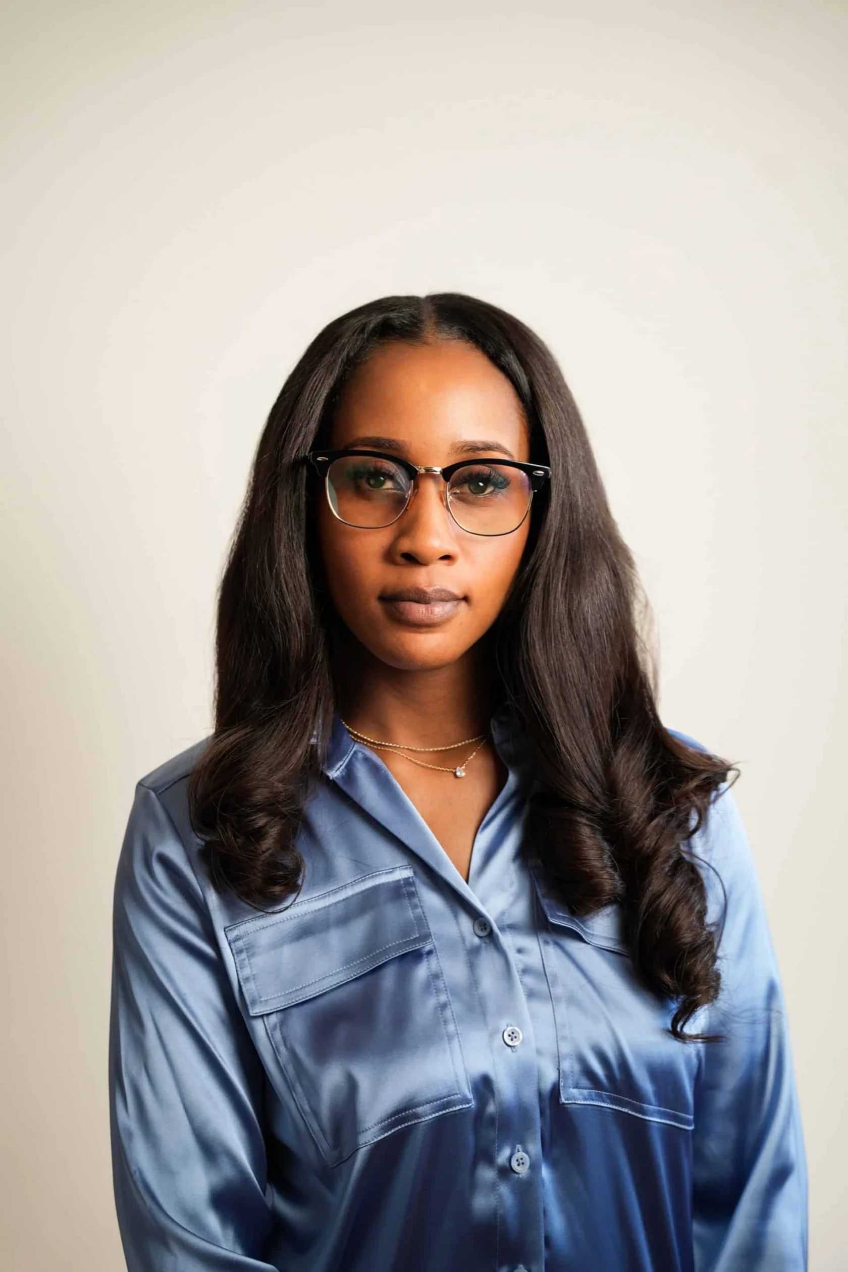 A woman with long, wavy hair and glasses wears a blue satin shirt and two necklaces, standing against a plain light background.