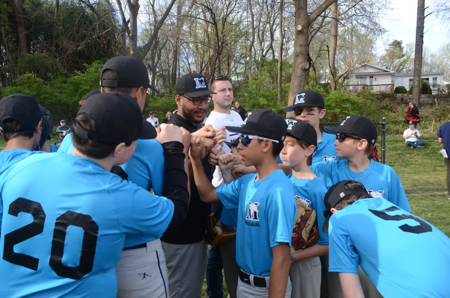 A youth baseball team in blue uniforms stands in a huddle with their coach, arms extended for a group cheer on a grassy field.