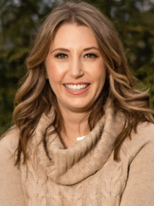 A woman with wavy light brown hair wearing a beige sweater is smiling outdoors with greenery in the background.