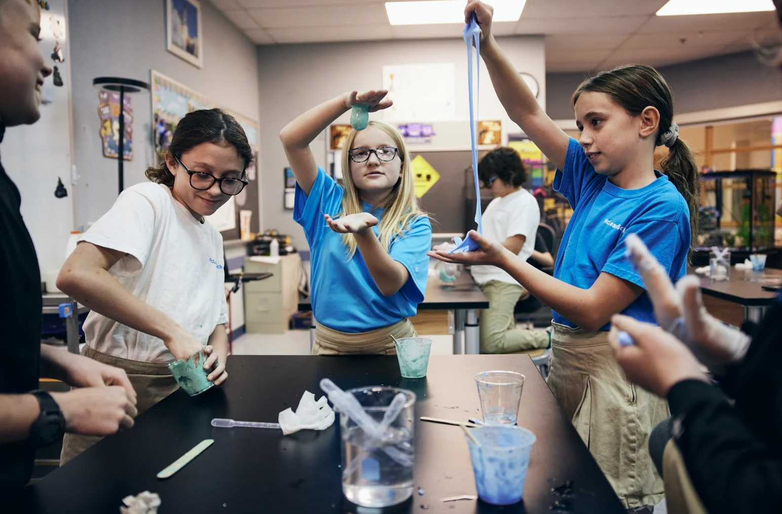 Four children in a science classroom are making slime with cups and stirrers, wearing casual clothes and looking focused on their activity.