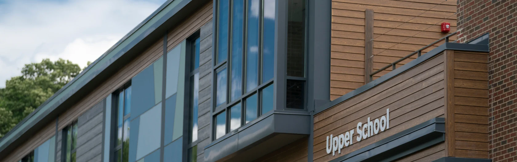 A modern building with wood paneling and large windows displays a sign that reads "Upper School" near the entrance.