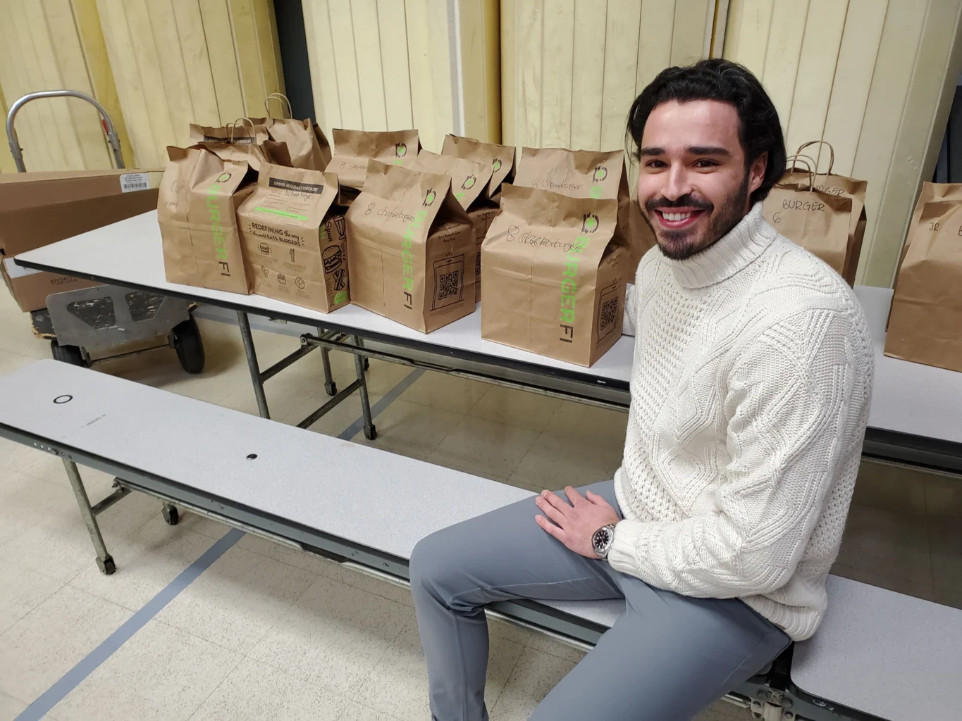 A man in a white sweater sits on a bench next to a row of paper grocery bags placed on a folding table indoors.