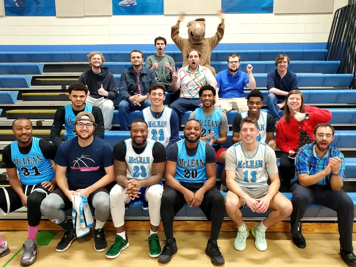 A basketball team in blue McLean jerseys sits in front of fans in bleachers; a person in a bear mascot costume stands in the back row, raising their arms.