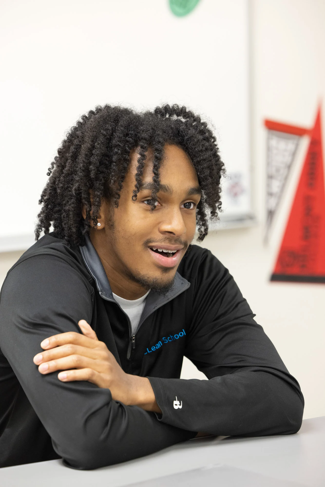 A young man with curly hair sits at a desk, leaning forward with his arms crossed and smiling. Classroom wall with banners is visible in the background.