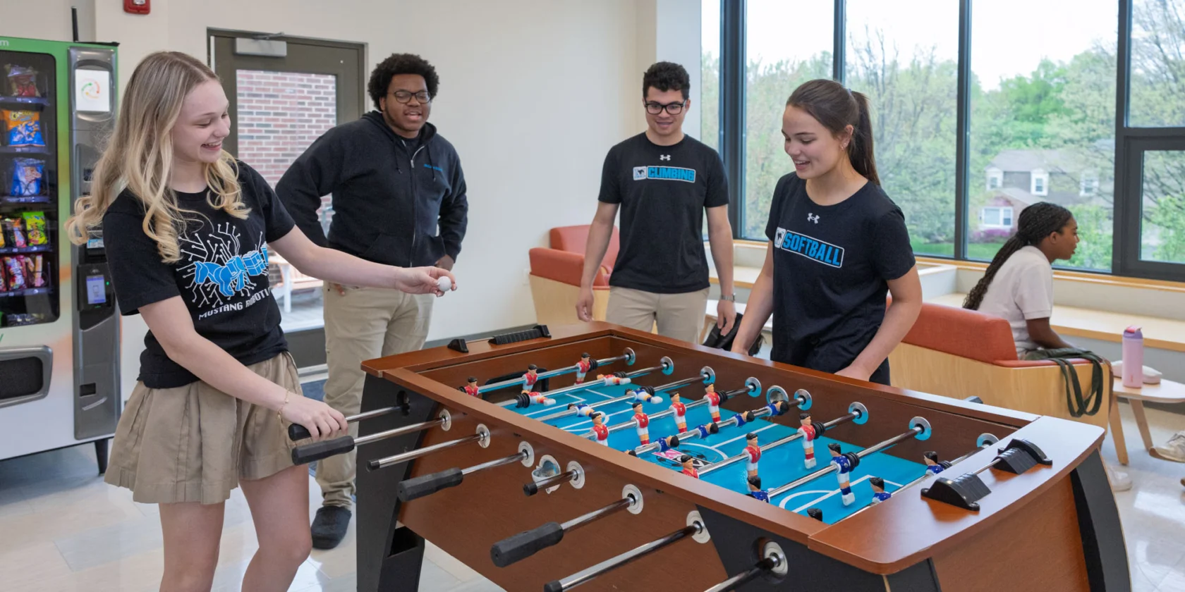 Four students play foosball in a common room while two others sit and talk on couches in the background.