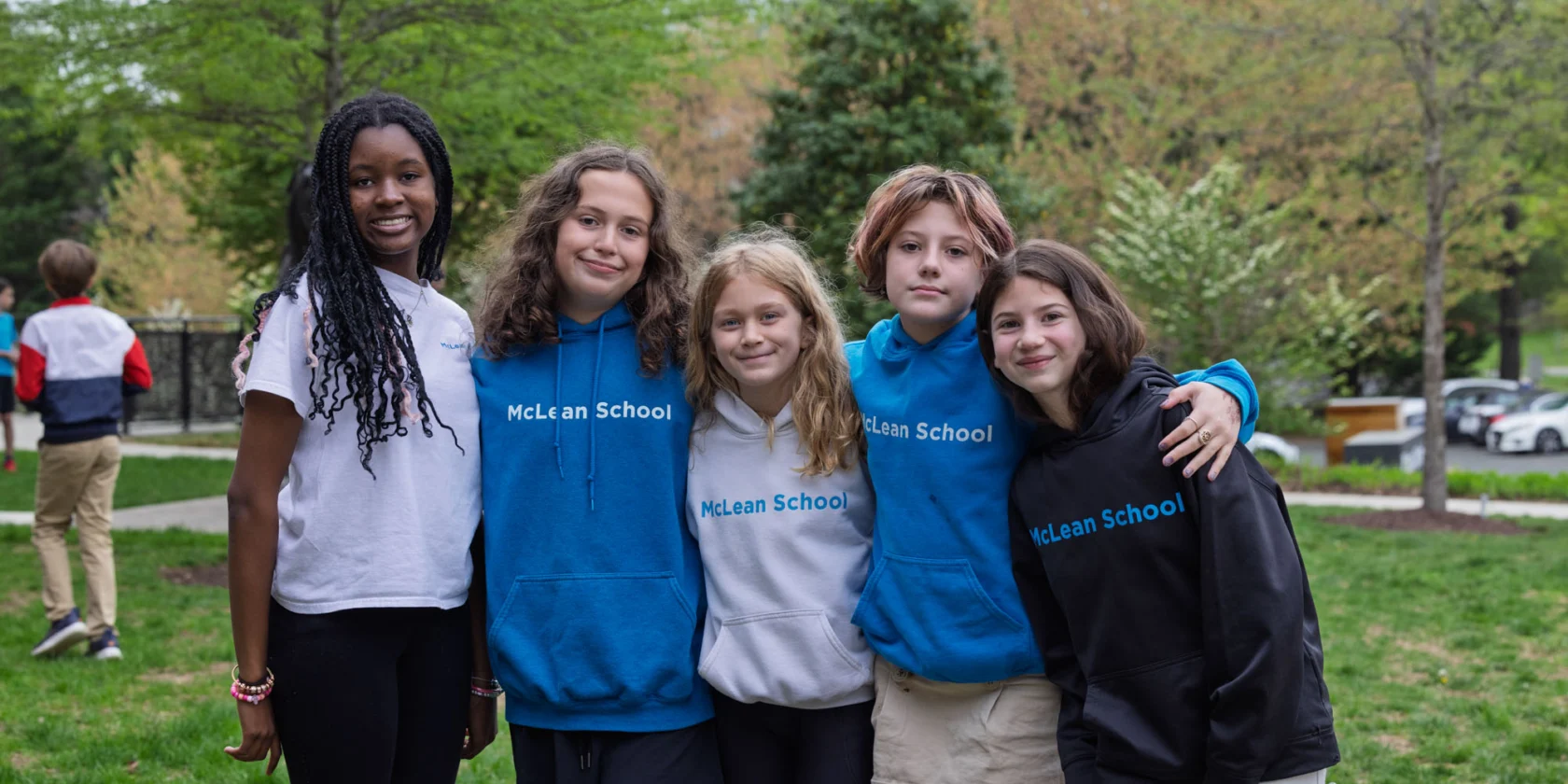 Five students stand together outside, three wearing blue "McLean School" hoodies, smiling at the camera. Trees and grass are visible in the background.