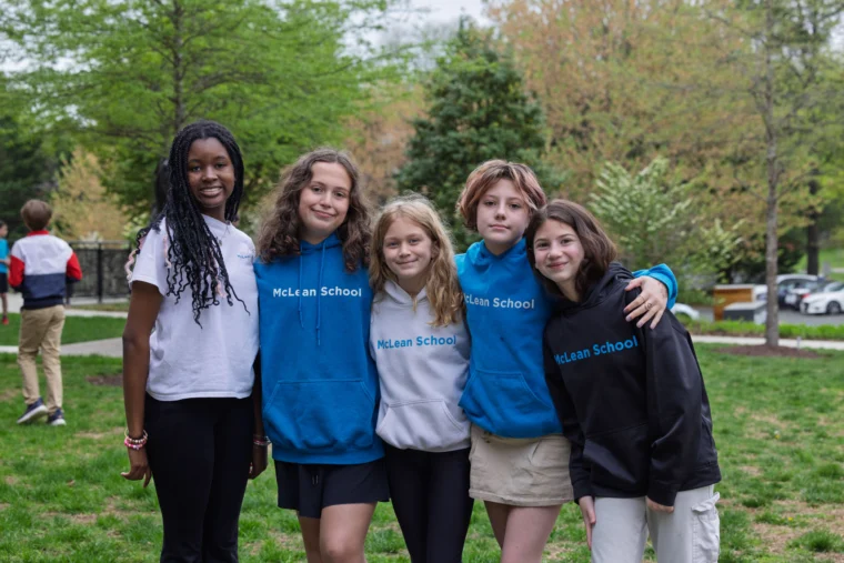 Five students stand together outside, three wearing blue "McLean School" hoodies, smiling at the camera. Trees and grass are visible in the background.