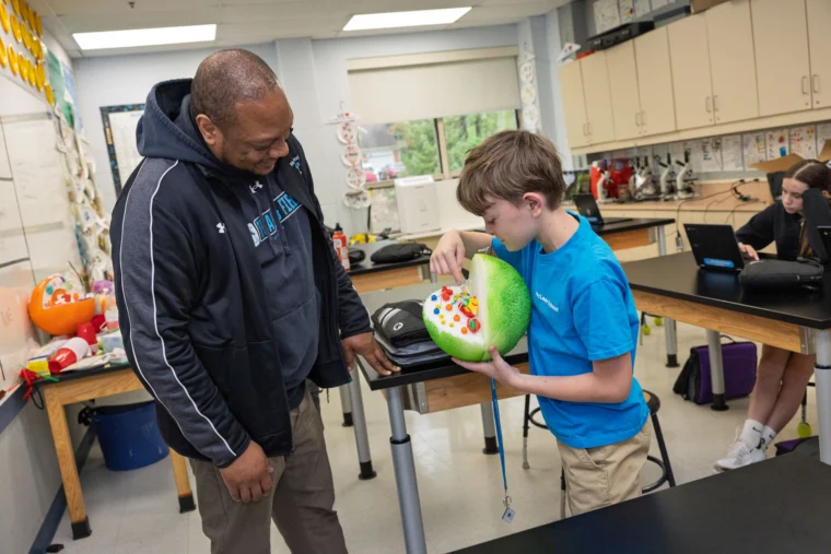 A student shows a teacher a large model of a cell in a science classroom while other students work at desks in the background.