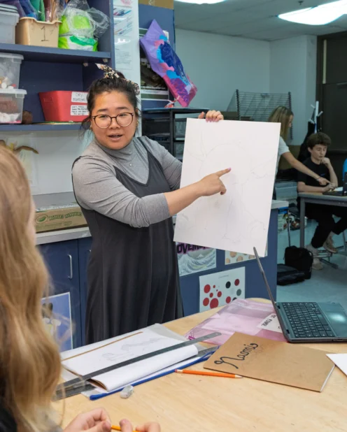 A teacher stands at a table showing a large sketch to students seated around her in an art classroom.