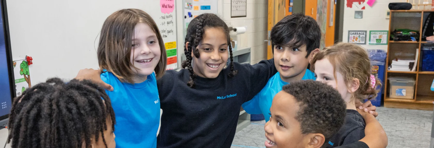 Six elementary-aged children stand in a circle with their arms around each other, smiling in a classroom setting.