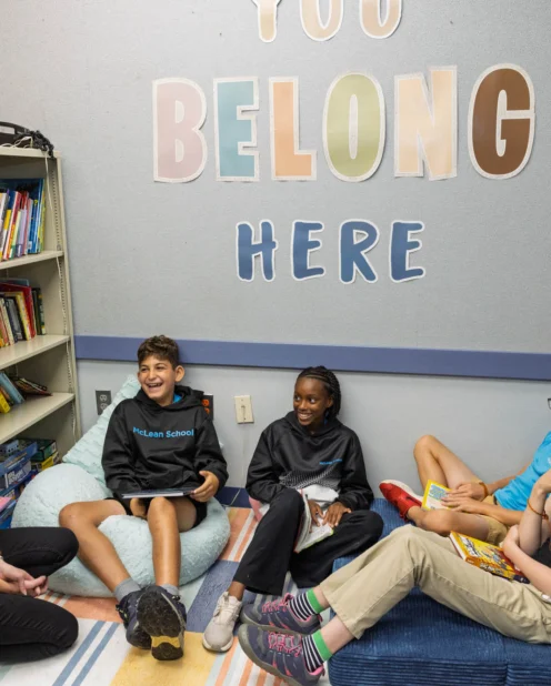 A teacher and four students sit together in a reading corner with books, under a wall sign that says "You Belong Here.