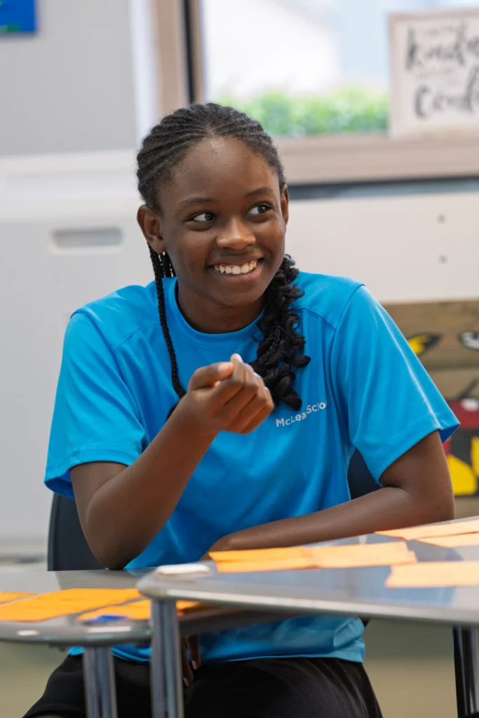 A girl in a bright blue shirt sits at a desk, smiling and gesturing with her hand in a classroom setting.