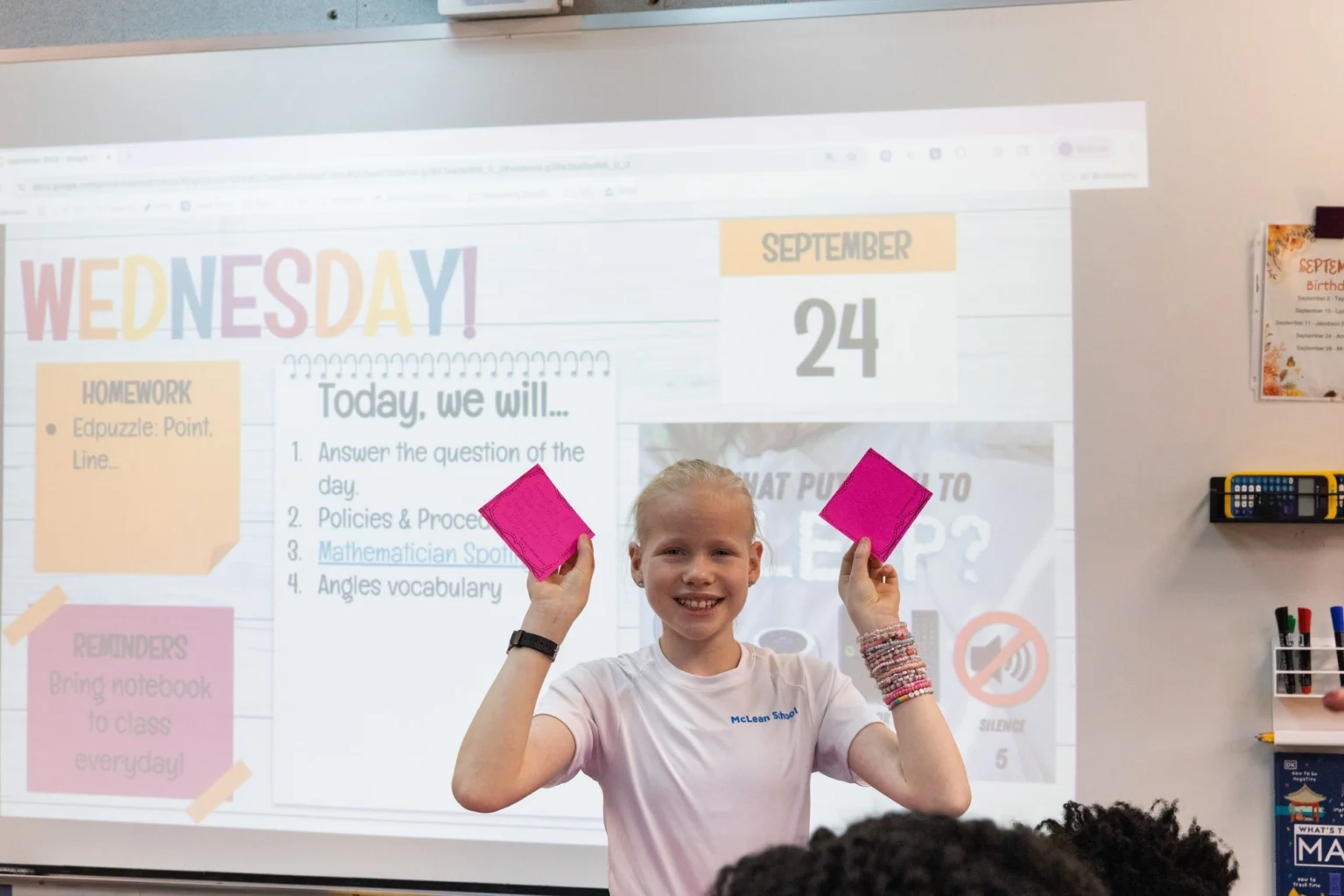 A student stands in front of a classroom whiteboard holding up two pink index cards, with the day's agenda and date, September 24, displayed on the screen behind them.