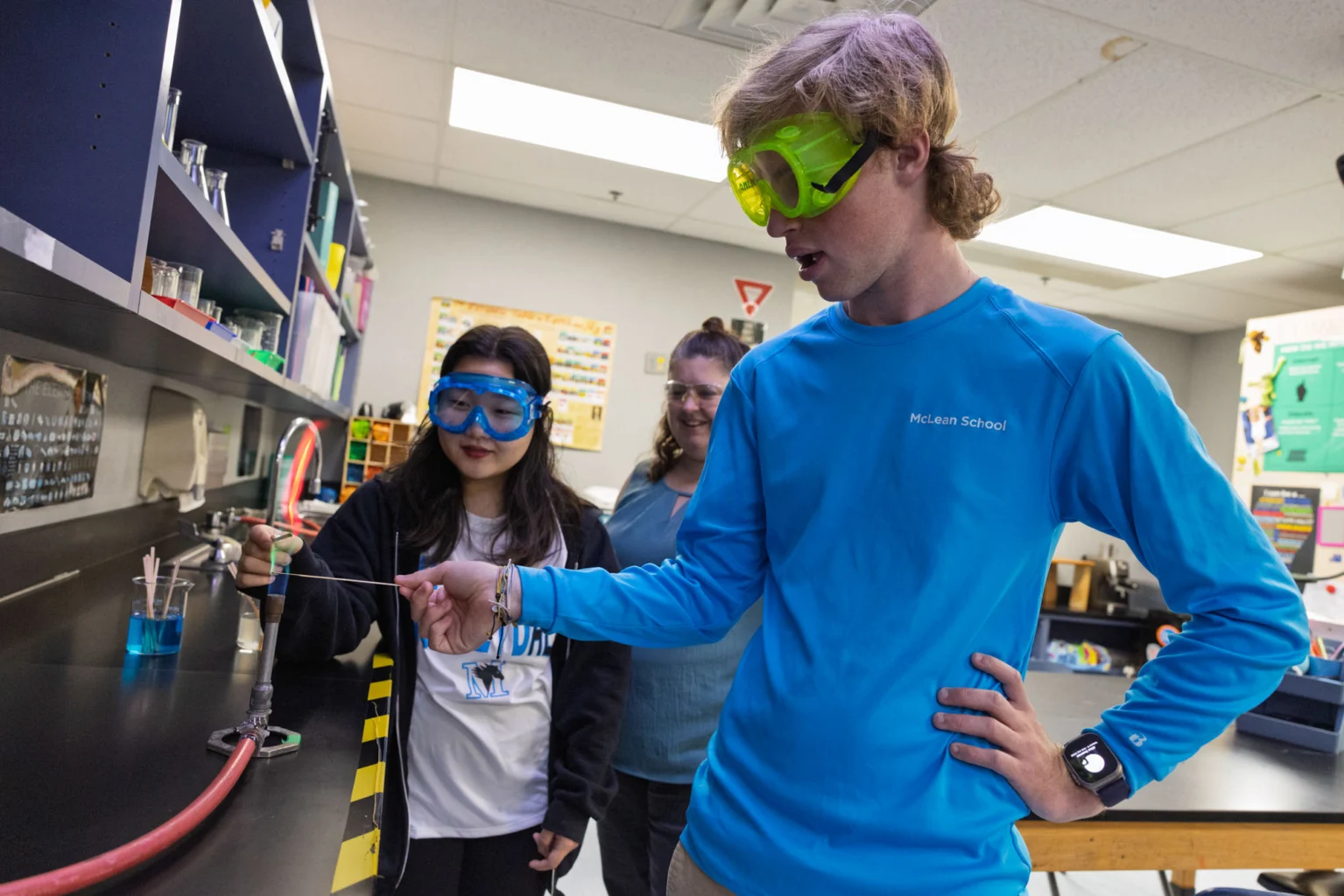 Three students wearing safety goggles conduct a science experiment at a lab bench, using beakers and a Bunsen burner in a classroom setting.