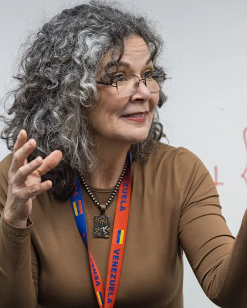 A woman with curly gray hair gestures while speaking in front of a whiteboard with writing on it. She wears glasses, a brown top, and a lanyard that says "Venezuela.