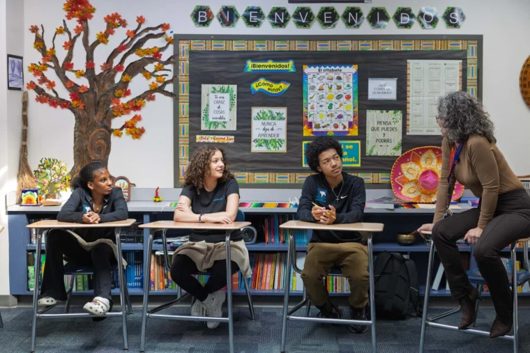 Three students sit at desks while a teacher leans on a desk and speaks to them in a colorful classroom with a "Bienvenidos" sign and educational posters on the wall.