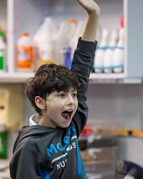A young boy in a classroom raises his hand enthusiastically, with his mouth open as if he is speaking or answering a question.