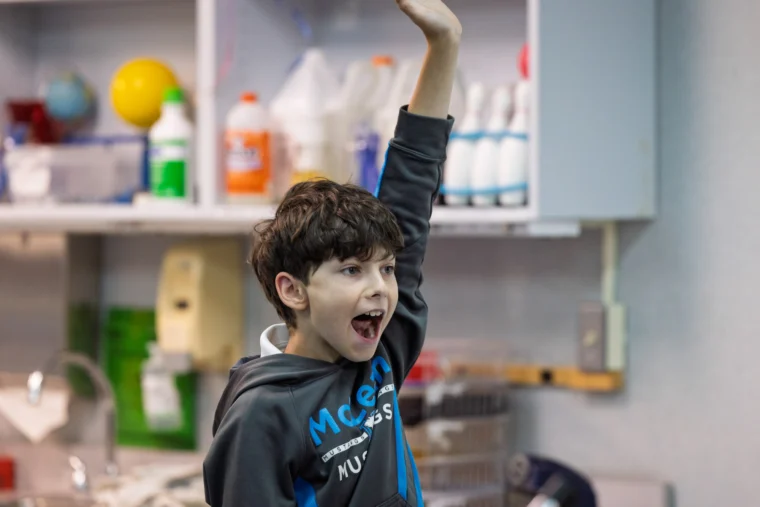 A young boy in a classroom raises his hand enthusiastically, with his mouth open as if he is speaking or answering a question.
