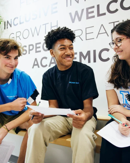Three students sit on a bench indoors, holding papers and pens, smiling and talking with each other in front of a wall with motivational words.