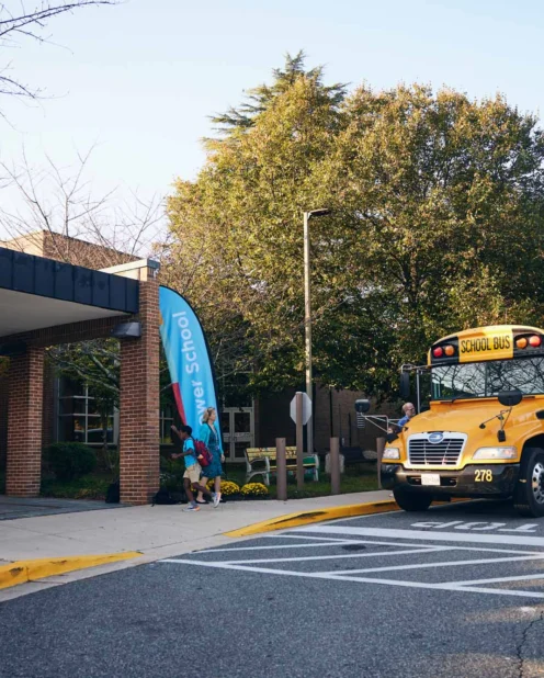 Two yellow school buses are parked outside a school building in the morning, with students walking toward the entrance and trees in the background.