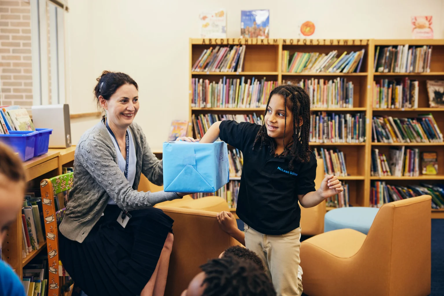 A teacher and a young student hold a blue box together in a library, surrounded by bookshelves and seated children.