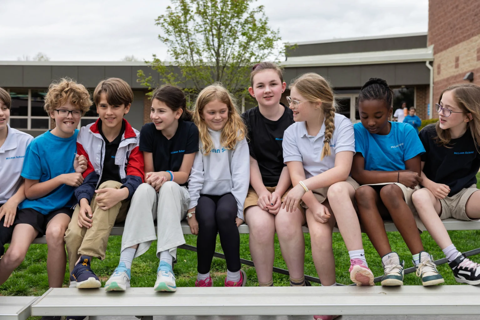 A group of school-age children sit on outdoor bleachers, smiling and talking together in front of a school building.