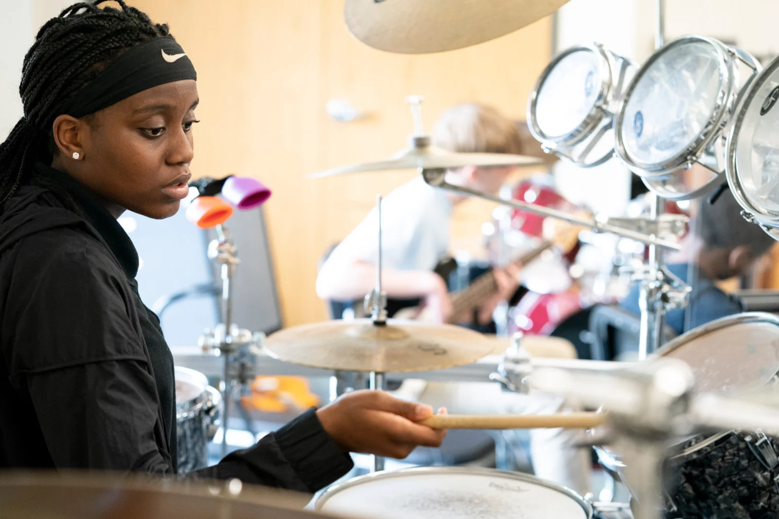 A young person in a black jacket and headband plays a drum set in a music room, with other musicians and instruments visible in the background.
