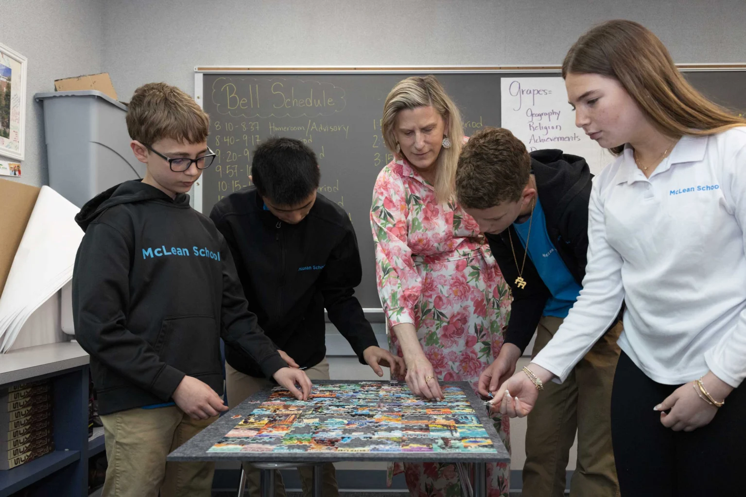 Four students and a teacher stand around a table, collaboratively working on assembling a jigsaw puzzle in a classroom.