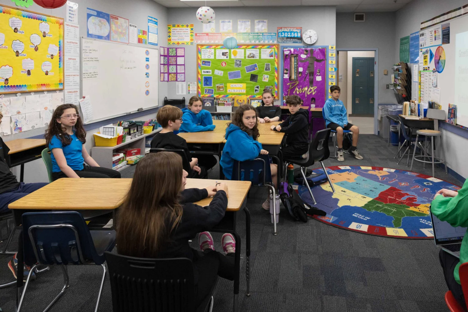 A classroom with students seated at desks and on chairs, facing a teacher. The room has colorful decorations, posters, and a rug with a map of the United States.