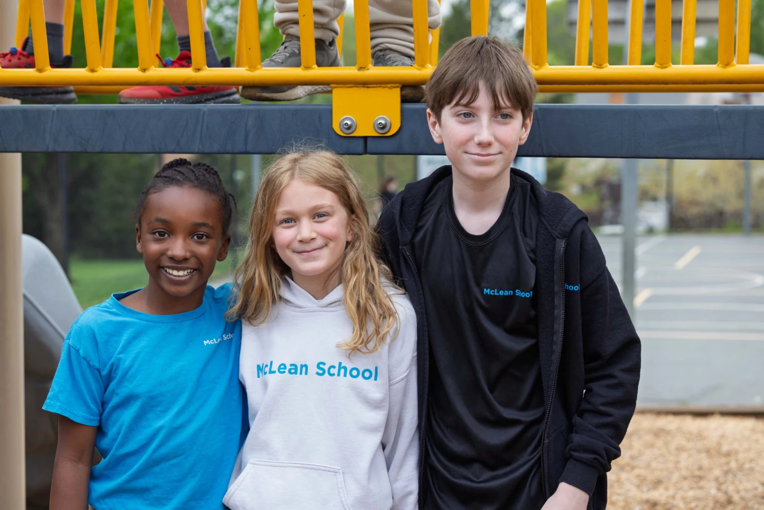 Three children stand together smiling under playground equipment, wearing shirts that say "McLean School" on a school playground.