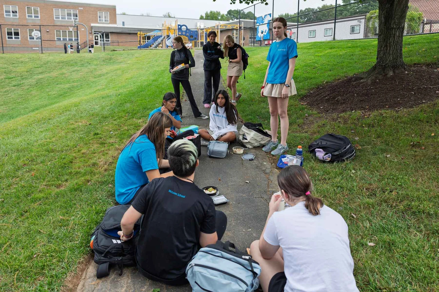 A group of students sit and stand on a paved path in a grassy area near a school building, eating lunch and talking. Backpacks and lunch containers are scattered nearby.