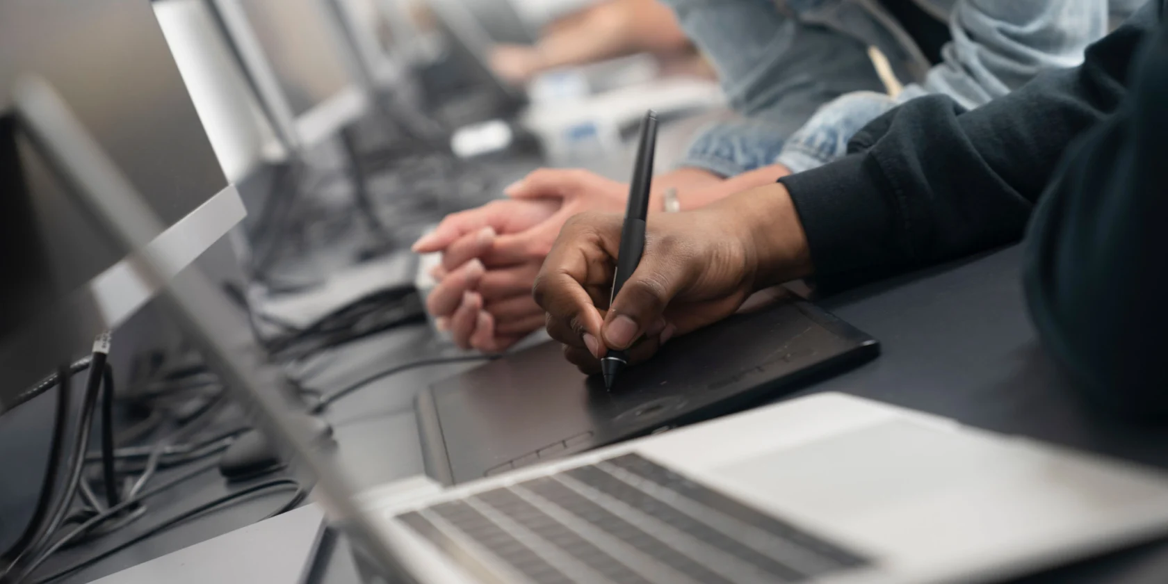 A person uses a stylus on a graphics tablet next to a laptop, while another person observes, in an office or classroom setting with multiple computers.