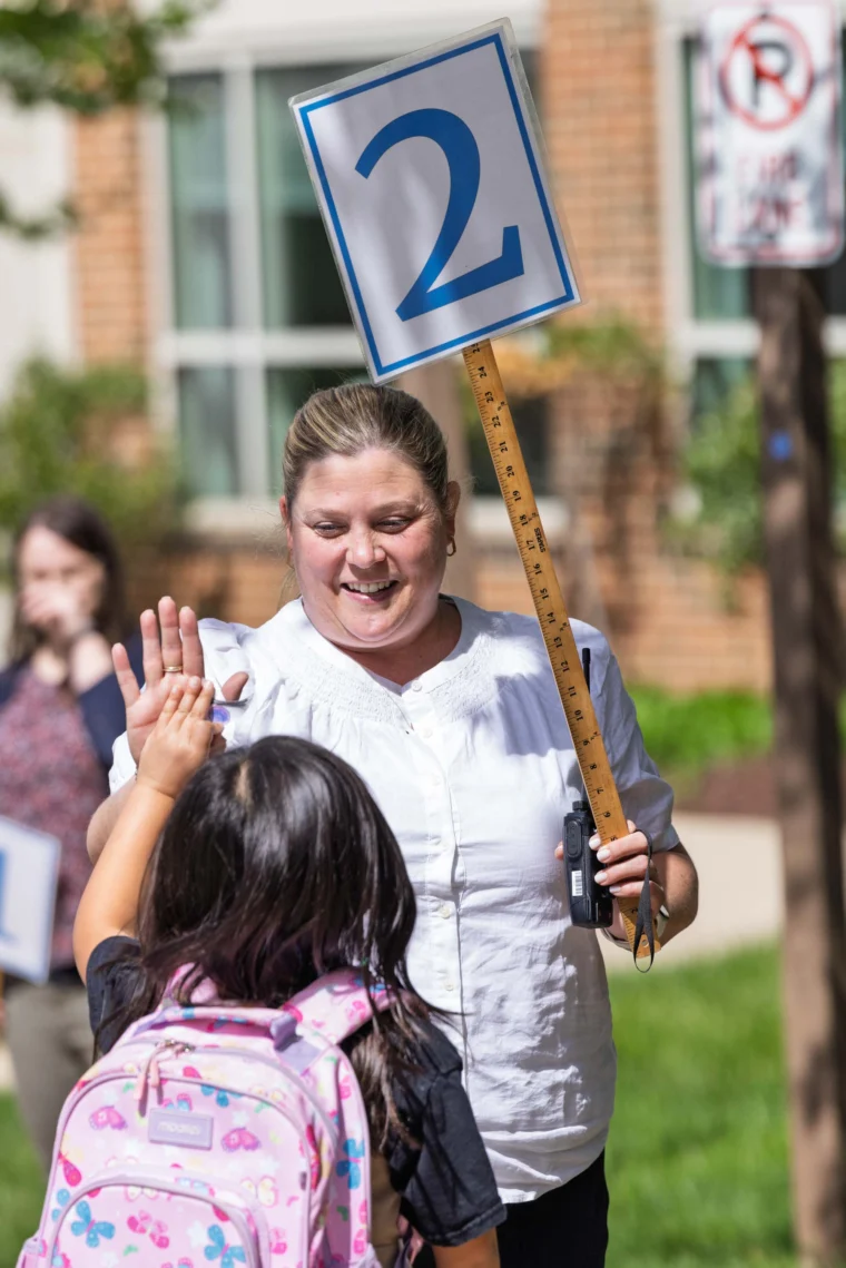 A woman holding a "2" sign on a ruler high-fives a child with a pink backpack outside a building.