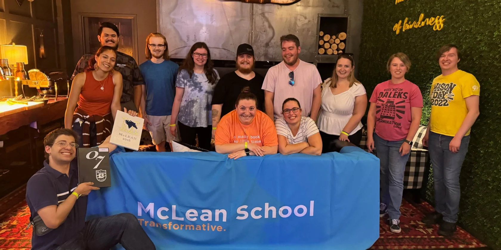 A group of people stand and sit behind a blue "McLean School" table inside a decorated room, smiling for the photo.