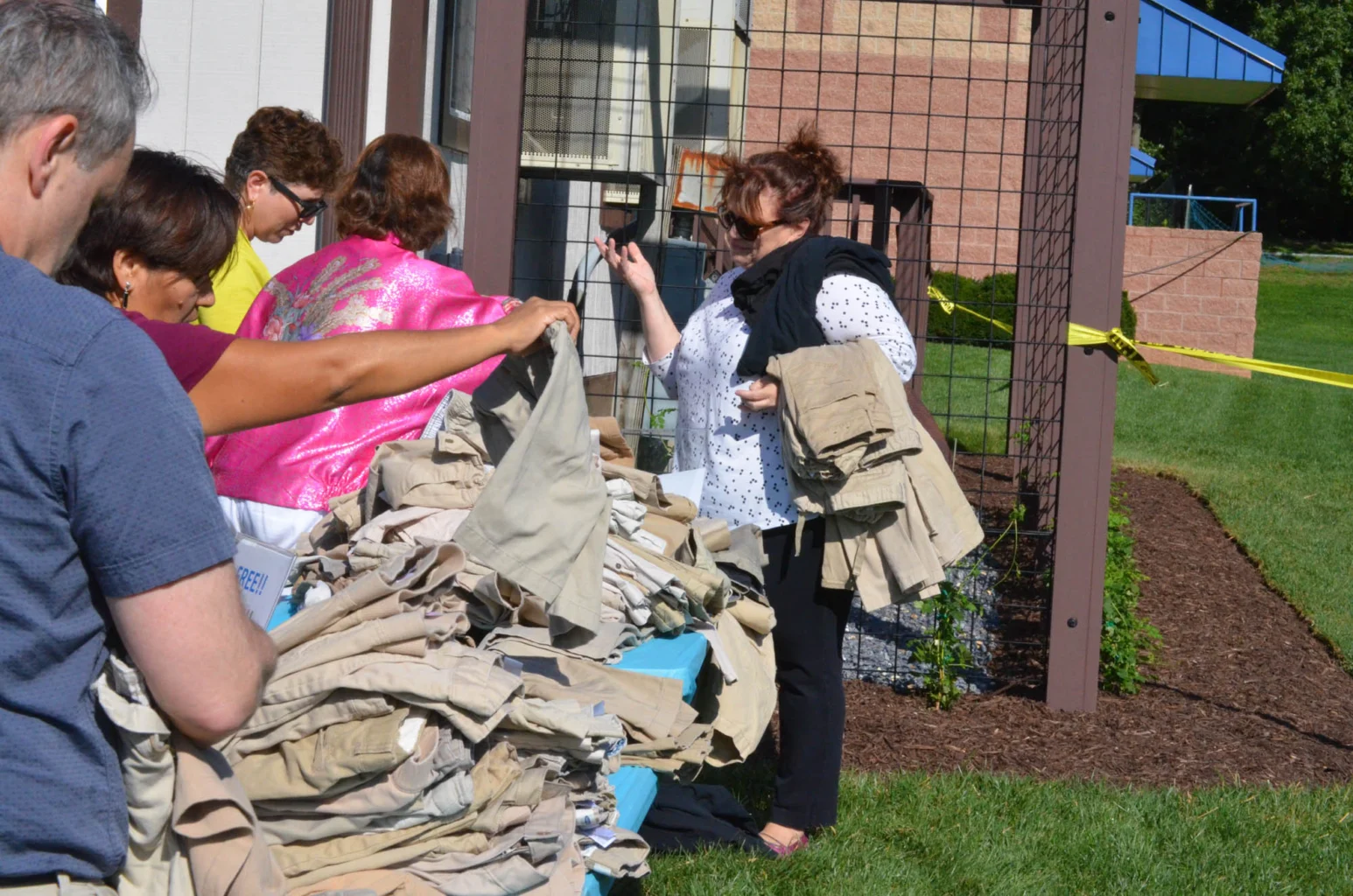 Several people stand outdoors near a large pile of khaki pants, sorting or collecting clothing items next to a building and a wire fence.