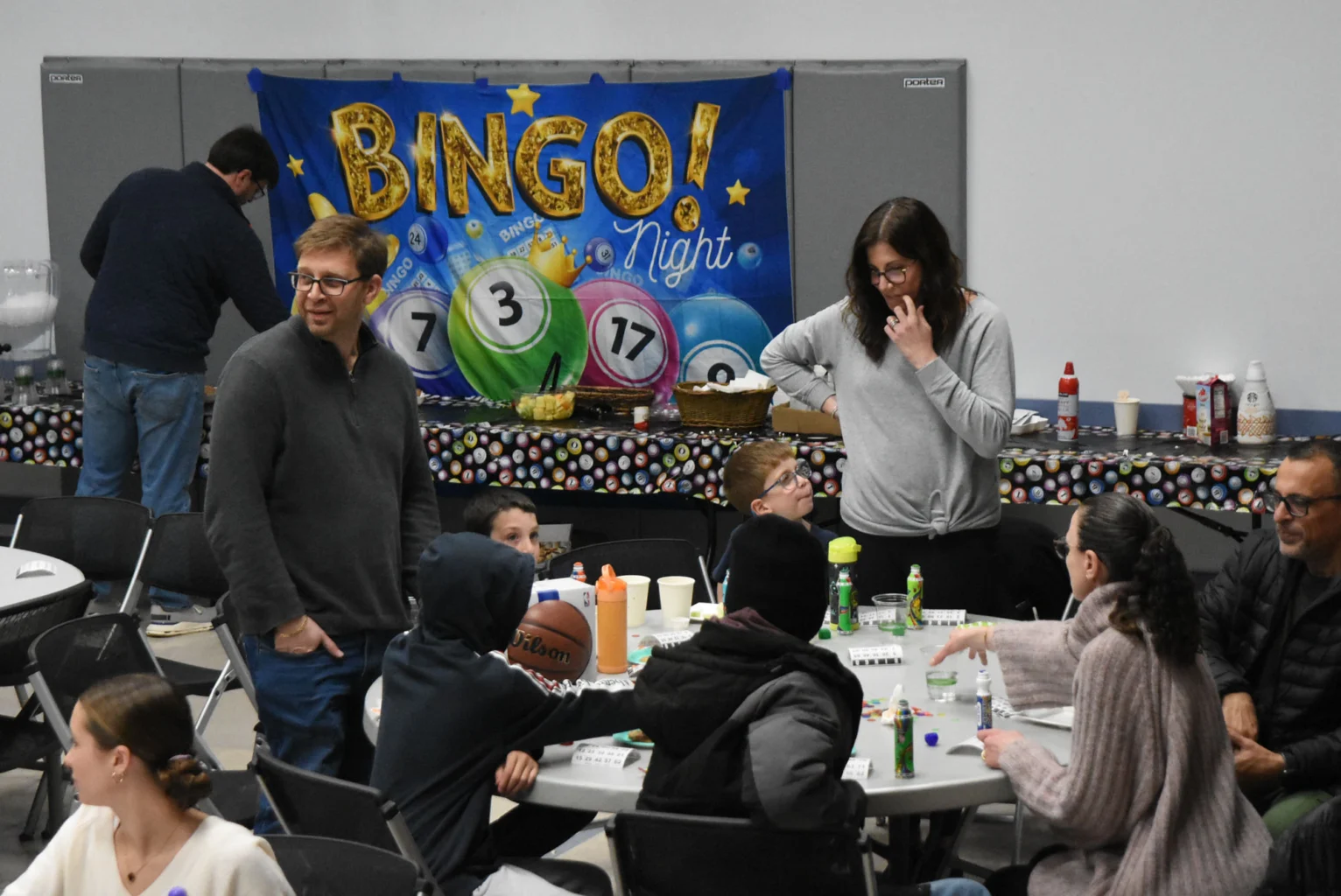 People sit and stand around tables during a bingo night event, with a colorful "Bingo Night" banner displayed in the background.