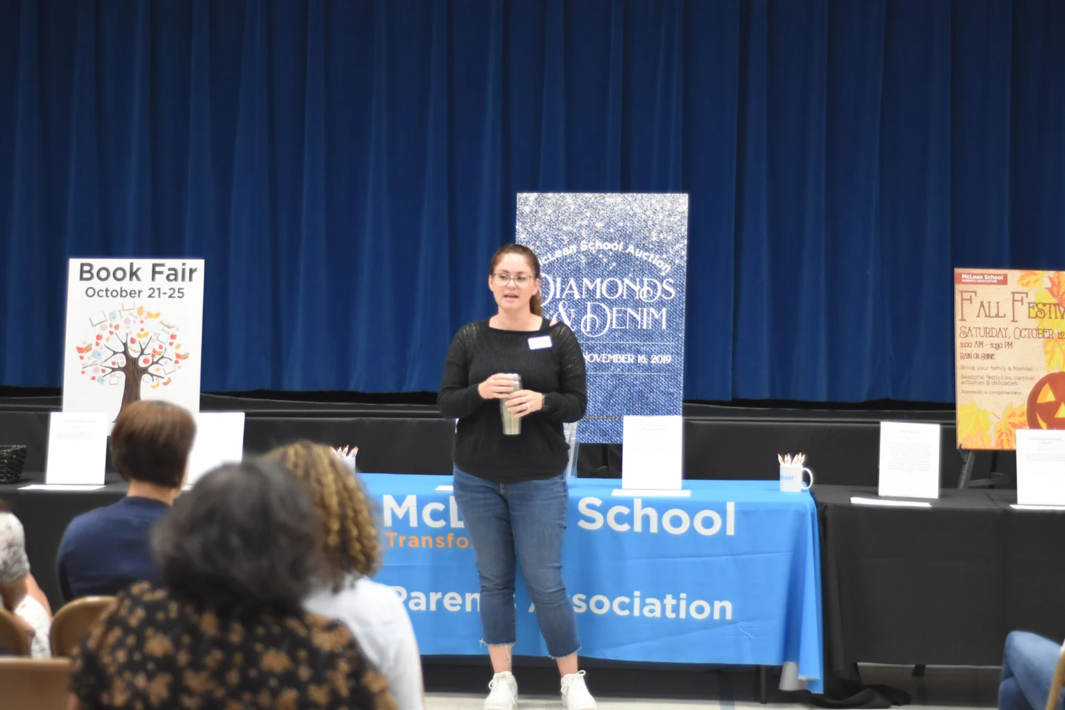 A woman speaks to an audience in a school gymnasium, standing behind a table with a blue McL School banner and event posters displayed in the background.