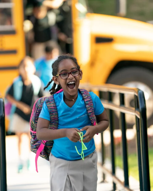 A young student with glasses and a backpack smiles while walking toward school, with a yellow school bus and other students in the background.