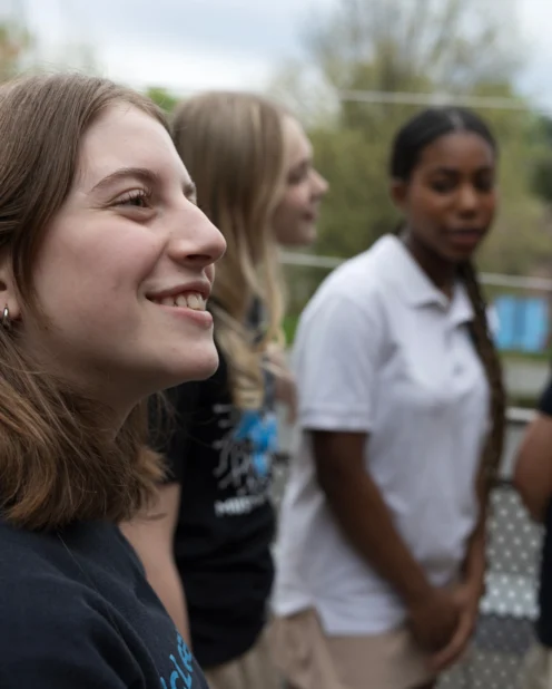 Four teenage girls stand outdoors, two in black shirts and two in white shirts, with trees and greenery in the background. One girl in the foreground is smiling.